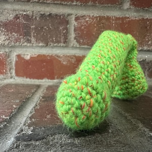 May include: A handmade, green crochet item with orange flecks. The textured yarn is formed into a rounded shape, possibly a decorative item or a small toy. The item is set against a brick background.