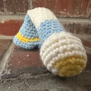 May include: A handmade crochet item in shades of white, light blue, and yellow. The item appears to be a decorative piece, possibly a toy or a small pillow, with a rounded shape and textured surface. The colors are soft and inviting.