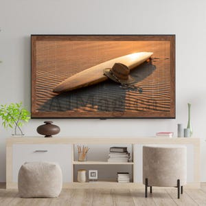 Puede incluir: Una obra de arte enmarcada representa una tabla de surf y un sombrero de paja sobre una superficie arenosa, bañados por la cálida luz del sol. La tabla de surf es de color marrón claro y el sombrero es de un tono marrón más oscuro. La obra está sobre un mueble blanco y marrón claro.