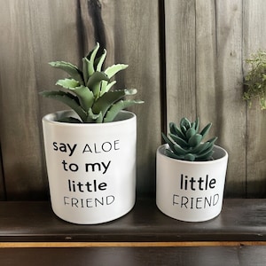 May include: Two white ceramic plant pots with black lettering. The larger pot has a succulent plant and says "say ALOE to my little FRIEND". The smaller pot has a succulent plant and says "little FRIEND".