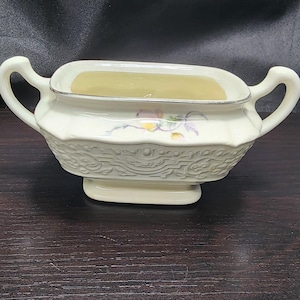 May include: Cream-colored ceramic sugar bowl with two handles and a decorative embossed pattern. The bowl has a rectangular opening with a silver rim and a small floral design. The sugar bowl sits on a rectangular base.