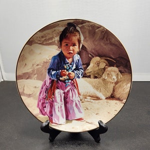 May include: A porcelain plate with a colorful image of a young girl in a pink dress and blue shirt holding a small doll. The girl is surrounded by two white goats in a rocky landscape.