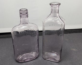 May include: Two antique, clear, lavender-colored glass bottles. One bottle is rectangular with the text "FULL PINT" embossed on the front. The other bottle is also rectangular with measurement markings on the side.