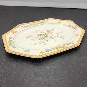 May include: An oval serving platter with a cream-colored surface and a floral design. The platter has a yellow and orange border with a scalloped edge. The floral design includes yellow, blue, and red flowers.