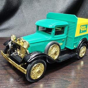 May include: A vintage-style toy truck in green, gold, black, and yellow. The truck features gold-colored wheels and trim, with a yellow cargo area. The text "Green Grocer" is visible on the side of the cargo area. The truck is a classic collectible.