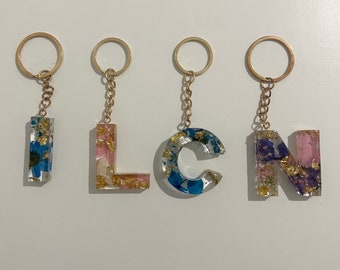 Puede incluir: Cinco llaveros dorados con dijes de letras de resina. Las letras deletrean "ILCN". Cada dije de letra está lleno de flores prensadas y pan de oro.