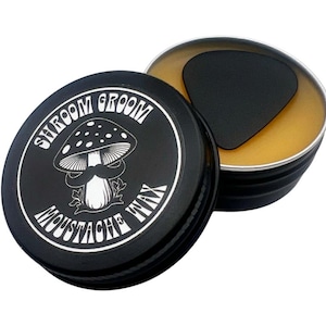 Peut inclure: Une boîte en métal noire avec une étiquette blanche qui dit "Shroom Groom Moustache Wax". L'étiquette représente un champignon de dessin animé avec une moustache. La boîte est ouverte et la cire à l'intérieur est de couleur jaune clair.