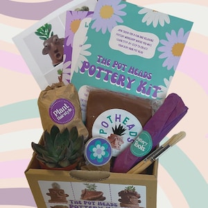 May include: A pottery kit in a cardboard box, including a turquoise and white booklet with the text "The Pot Heads Pottery Kit." The kit contains clay, paintbrushes, a succulent plant, and a "Plant Hairstyle" bag. Measurements are in cm and grams.