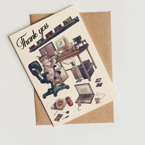 May include: A greeting card with the words "Thank you" in script. The card features a whimsical illustration of a cat wearing glasses, sitting in an office chair, surrounded by computers, a pizza, and a laptop. The card is on a brown envelope.