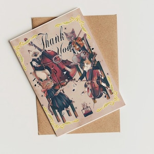 May include: A greeting card with a vintage design featuring cats playing musical instruments. The card has the words "Thank You" in elegant script. The card is on top of a brown envelope, with a decorative yellow border.