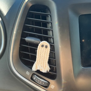 May include: A white crocheted ghost car air freshener with black eyes hanging from a car's air vent.
