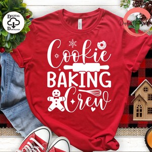 Może przedstawiać: Czerwona koszulka z białym napisem "Cookie Baking Crew". Wzór zawiera piernikowego ludzika, wałek do ciasta, płatek śniegu, pączek i trzepaczkę.