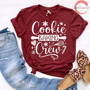 May include: A maroon t-shirt with white text that reads "Cookie Baking Crew" and includes a snowflake, rolling pin, whisk, and candy cane graphic.