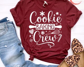 Może przedstawiać: Bordowy t-shirt z białym napisem "Cookie Baking Crew" i grafiką przedstawiającą płatek śniegu, wałek do ciasta, trzepaczkę i laskę cukrową.
