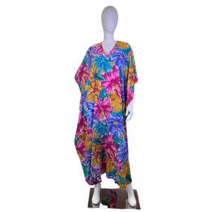 Puede incluir: Un vibrante vestido caftán con estampado floral. El vestido presenta un cuello en V y mangas anchas. La tela está adornada con grandes flores coloridas en tonos rosa, azul, amarillo y verde. El vestido se exhibe en un maniquí.