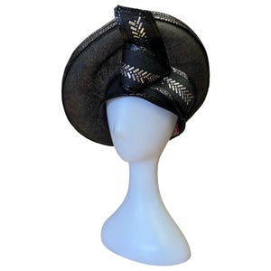 May include: A black hat with a wide brim and a decorative bow. The bow and trim feature silver embellishments. The hat is displayed on a white mannequin head, showcasing its shape and design. The hat appears to be made of a textured material.