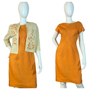 Könnte beinhalten: Zwei Vintage-Kleider auf Schaufensterpuppen. Eines ist ein leuchtend orangefarbenes Etuikleid. Das andere ist ein passendes orangefarbenes Kleid mit einer cremefarbenen Jacke mit floralen Ausschnitten. Die Kleider sind knielang und scheinen aus leichtem Stoff zu sein.