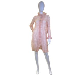 May include: A pale pink lace duster coat featuring long sleeves with ruffled cuffs, a high ruffled collar, and a vertical ruffled placket down the front. The garment is secured with two circular, silver-toned decorative buttons with clear crystals.