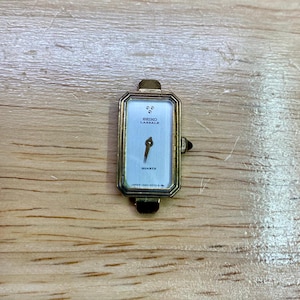 Pode incluir: Um relógio Seiko Lassale Quartz retangular com caixa dourada e mostrador azul claro. O mostrador do relógio apresenta ponteiros pretos e o texto "SEIKO LASSALE QUARTZ". O relógio está sobre uma superfície de madeira.