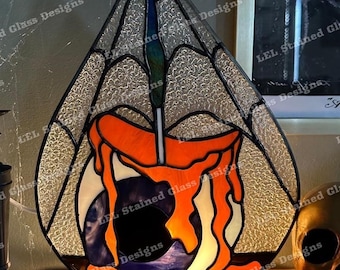 Puede incluir: Una lámpara de vidrio tintado con forma de lágrima con un diseño de telaraña y una vela que gotea en su interior. La lámpara está encendida y proyecta un cálido resplandor. La lámpara está sobre una base de madera y hay una calavera dorada en el fondo.