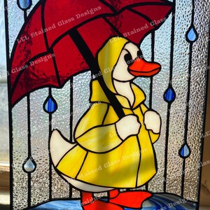 May include: A stained glass window depicting a yellow duck wearing a raincoat and holding a red umbrella. The duck is standing in a puddle of water with raindrops falling around it. The window is framed in black.