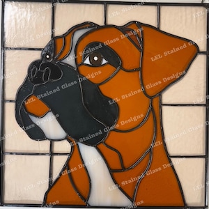 Puede incluir: Ventana de vitral que representa un perro boxer marrón y negro con una mancha blanca en el pecho. La cabeza del perro está girada hacia un lado y sus ojos están cerrados. El fondo es de color beige claro.