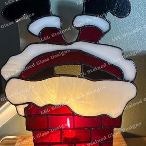 May include: A stained glass depiction of Santa Claus stuck in a chimney. The chimney is made of red brick and the Santa Claus is wearing a red suit with white trim. The glass is illuminated from behind, creating a warm glow.