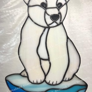 May include: Stained glass polar bear on an iceberg. The bear is white with black detailing, and the iceberg has blue hues. The design uses black lines and includes a hanging chain for display. The piece is made of glass.