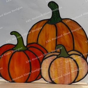 May include: A stained glass pumpkin trio in orange and yellow with green stems. The pumpkins are grouped together, with different sizes and shapes. The glass has a textured look, and the design is outlined in black.