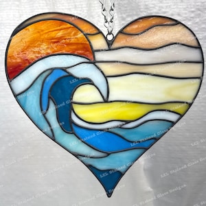 May include: A heart-shaped stained glass suncatcher featuring a sunset and ocean wave design. The artwork uses shades of orange, yellow, blue, and white. It is suspended by a silver chain. The piece is a decorative item.