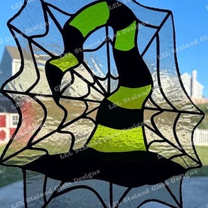 May include: A stained glass window hanging, featuring a witch's hat with black and green stripes, set against a spiderweb design. The text "LEL Stained Glass Designs" appears multiple times around the border.
