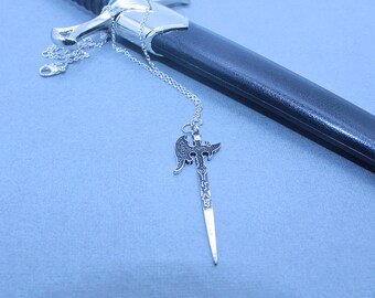 May include: A silver necklace with a sword pendant. The sword has a detailed design and a pointed tip. The necklace is lying on a blue surface next to a black leather sword scabbard.