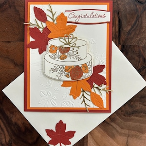 May include: A rectangular greeting card with a white embossed background featuring a two-tier cake illustration decorated with orange and red floral patterns. The card includes die-cut red and orange maple leaves and the text Congratulations.