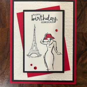 May include: A handmade birthday card with a red and black border. The card features a sketch of the Eiffel Tower and a woman in a red hat, with the words "Happy Birthday, Gorgeous."