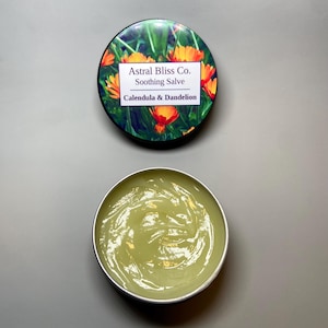May include: A metal tin of soothing salve with the lid off, showing a light green, creamy substance. The lid has a floral design with the text "Astral Bliss Co. Soothing Salve Calendula & Dandelion."