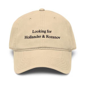 Peut inclure: Casquette de baseball beige avec le texte "Looking for Hollander & Rozanov" en noir. La casquette a une visière incurvée et un bouton sur le dessus. Le chapeau est en tissu doux.