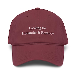 May include: A maroon baseball cap with the text "Looking for Hollander & Rozanov" in white. The cap has a curved brim and a button on top. The hat is made of a soft fabric.