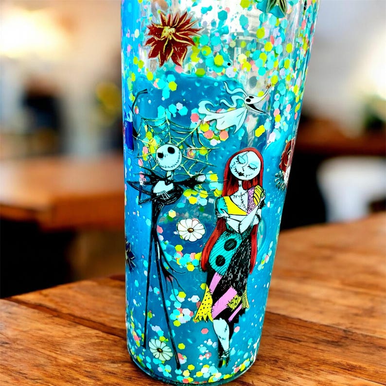 May include: A tall, clear tumbler with a blue background and floating glitter. The tumbler features illustrations of Jack Skellington and Sally from The Nightmare Before Christmas. The design includes flowers and other decorative elements.