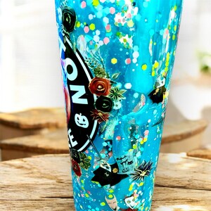 May include: A tall, clear tumbler filled with blue liquid and colorful glitter. The tumbler features a black and white sticker with text and floral accents. Decorative elements include small bats and a cartoon character.