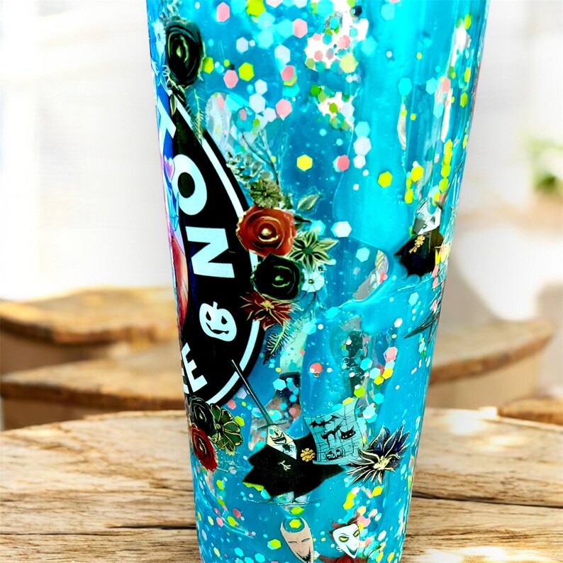 May include: A tall, clear tumbler filled with blue liquid and colorful glitter. The tumbler features a black and white sticker with text and floral accents. Decorative elements include small bats and a cartoon character.