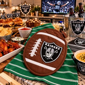 May include: A brown, oval-shaped oven mitt designed to look like a football, featuring white laces and the Raiders team logo in the center. The mitt is placed on a green and white striped kitchen towel, surrounded by various game day food items.