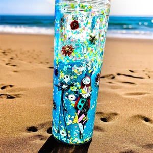 May include: A tall, clear tumbler with a blue background and a design featuring Jack Skellington and Sally from The Nightmare Before Christmas. The tumbler is filled with glitter and decorative elements, with a clear lid.