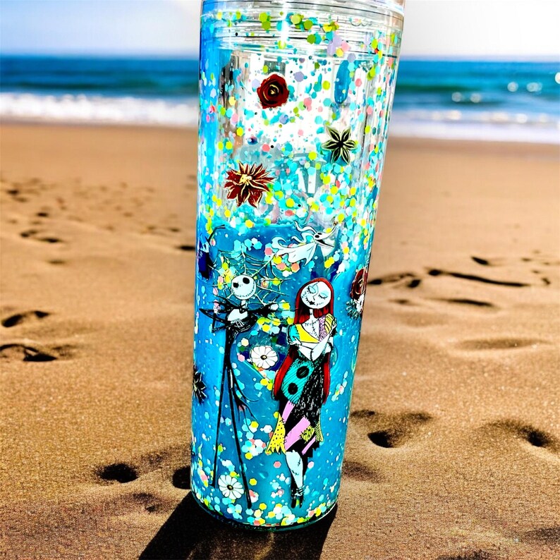 May include: A tall, clear tumbler with a blue background and a design featuring Jack Skellington and Sally from The Nightmare Before Christmas. The tumbler is filled with glitter and decorative elements, with a clear lid.