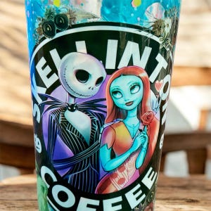 May include: A clear tumbler with a black and white graphic of Jack Skellington and Sally from The Nightmare Before Christmas. The tumbler has the words "KELLY INTO COFFEE" in white and green. The background is a light blue with glitter.