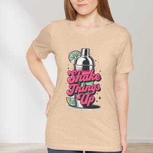 May include: A tan t-shirt with a graphic of a cocktail shaker with a pink and green drink inside. The text "Shake Things Up" is printed in pink letters on the shirt.