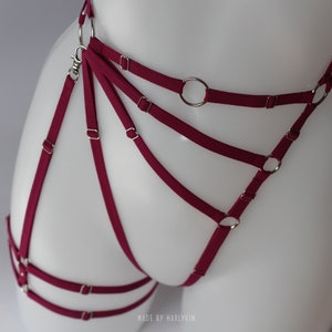 May include: Burgundy red lingerie harness with silver metal rings and clasps.  The harness has multiple straps that crisscross and connect to the rings.  "MADE BY HARLYKIN" is printed on the bottom of the image.