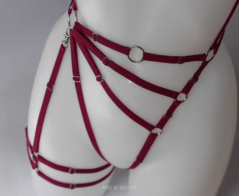 May include: Burgundy red lingerie harness with silver metal rings and clasps.  The harness has multiple straps that crisscross and connect to the rings.  "MADE BY HARLYKIN" is printed on the bottom of the image.