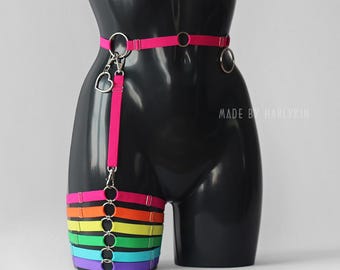 May include: A rainbow colored thigh harness with a pink strap and silver rings. The harness is made of elastic and has a heart-shaped charm attached to the strap. The text "MADE BY HARLYKIN" is visible on the mannequin.
