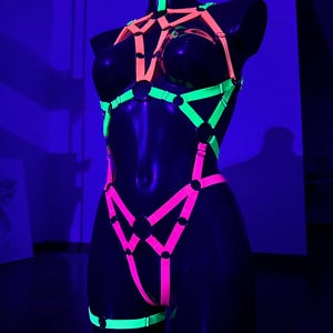 May include: A neon-colored body harness displayed on a black mannequin. The harness features bright pink, green, and orange straps with metal rings. The text "MADE BY HARLYKIN" is visible at the bottom. The overall design is geometric and eye-catching.