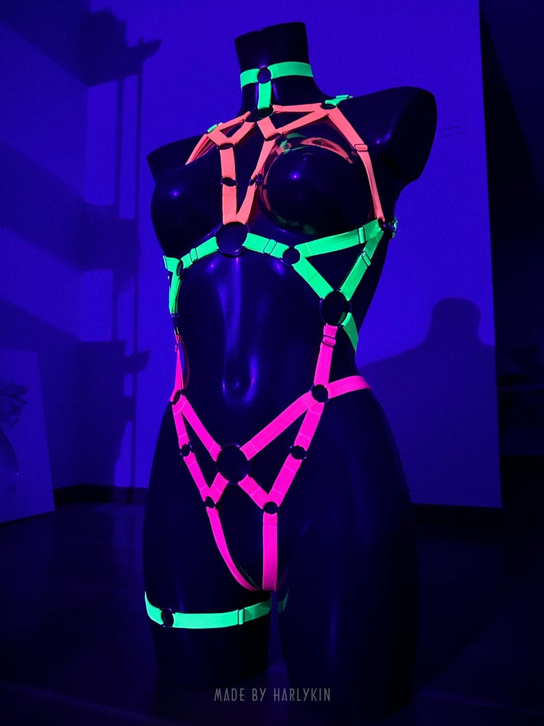 May include: A neon-colored body harness displayed on a black mannequin. The harness features bright pink, green, and orange straps with metal rings. The text "MADE BY HARLYKIN" is visible at the bottom. The overall design is geometric and eye-catching.